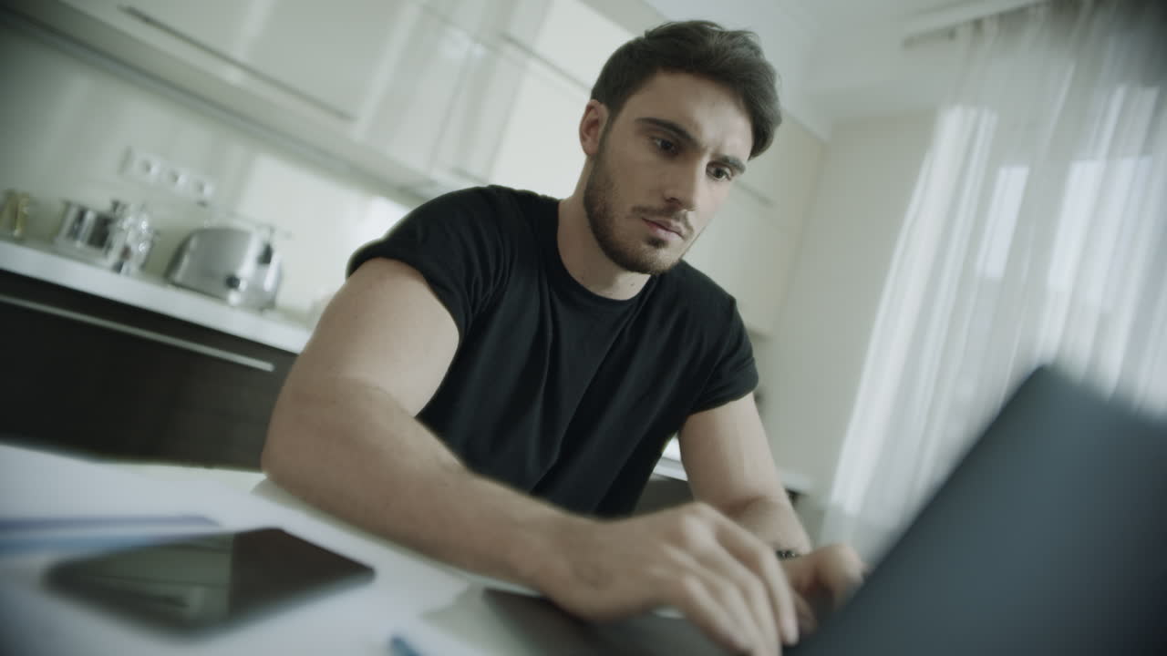 Uomo serio che digita al computer portatile a casa. Uomo d'affari che usa il computer in cucina