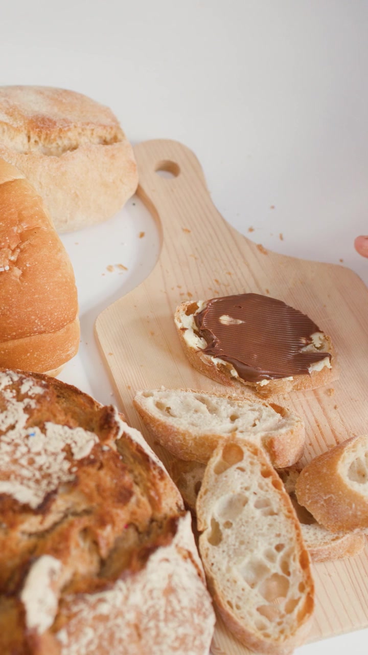 Hands spread chocolate cream on sliced bread amidst sourdough, baguettes, and croissants on wood.