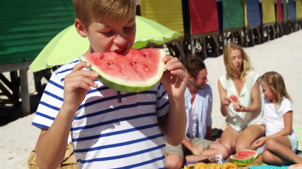 niño comiendo sandía mientras la familia está sentada en el fondo en la playa