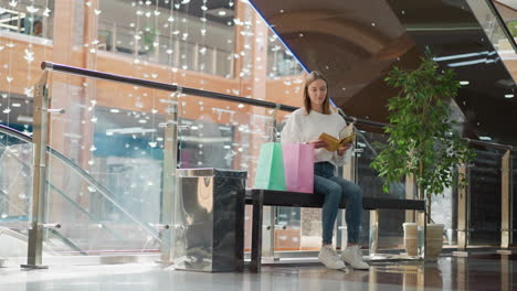 young woman seated on bench in modern mall opens yellow book thoughtfully, surrounded by shopping bags and elegant decor with glass railings, decorative plants, and warm light accents