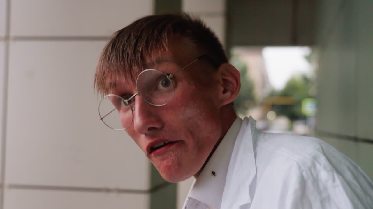 Young man in white coat wearing glasses standing on porch, chewing mouth while sipping unusual green drink from bottle, taking break in urban outdoor setting with thoughtful expression