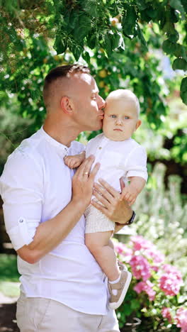 happy caucasian father in white clothes holding a little baby boy. loving dad kisses his son and attracts his attention. garden at backdrop. vertical video.