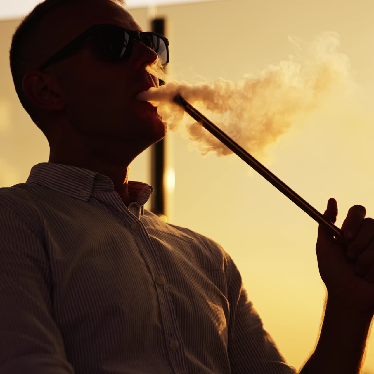 Man in dark glasses sitting at sunset holding hookah pipe. Relaxing male making thick smoke from nargileh. Close up