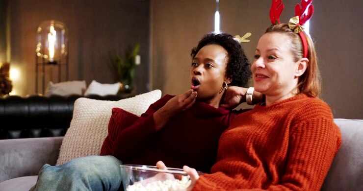 Amiche multietniche sedute sul divano che condividono popcorn in una ciotola di vetro, reagendo a un film di Natale.