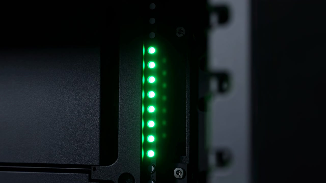 Close-up of dark server hardware with a vertical row of glowing green indicator lights, blurred equipment in the foreground, and low-key industrial lighting