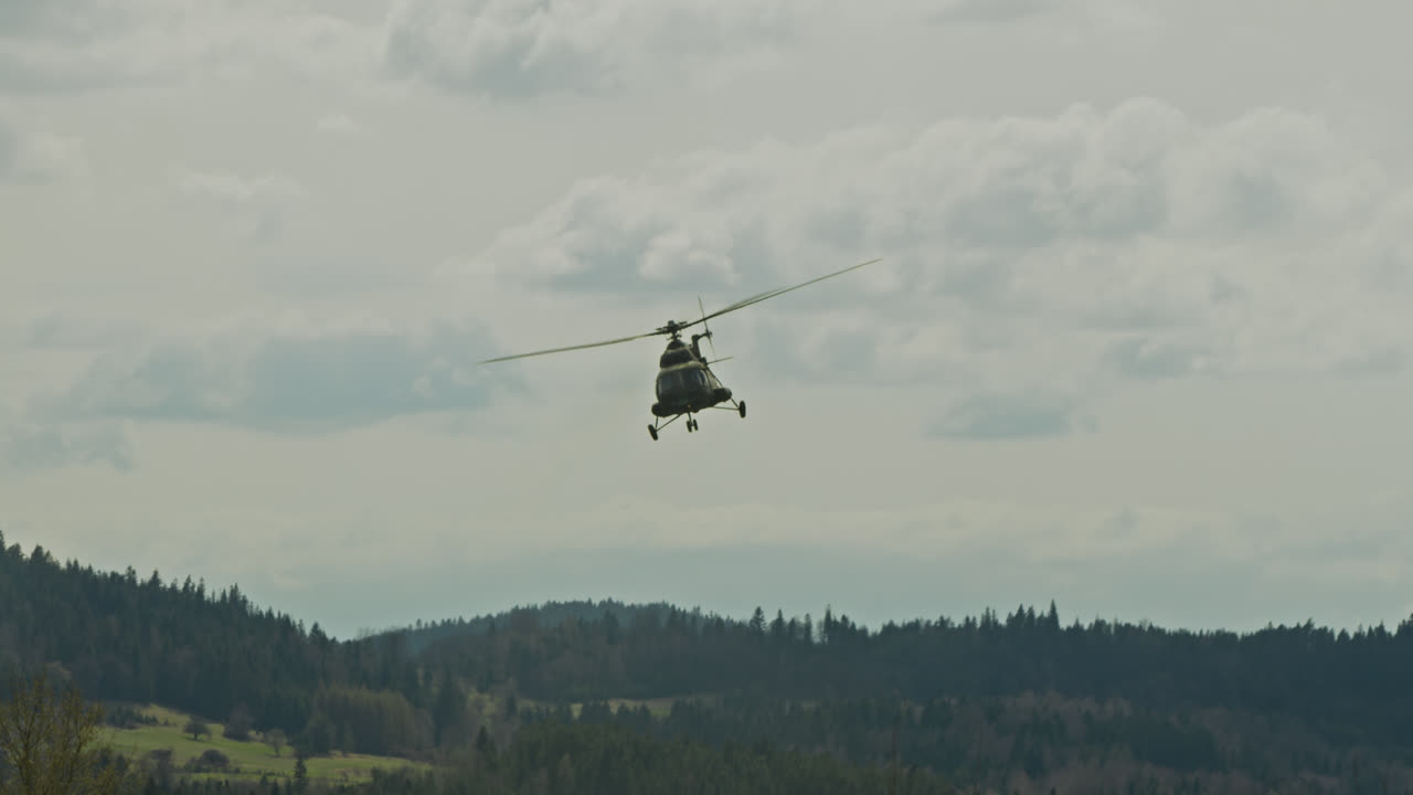 A military-style helicopter flies over dense coniferous forest with overcast skies above, creating a dynamic contrast between nature and machinery. Captured in a mountainous region during a day.