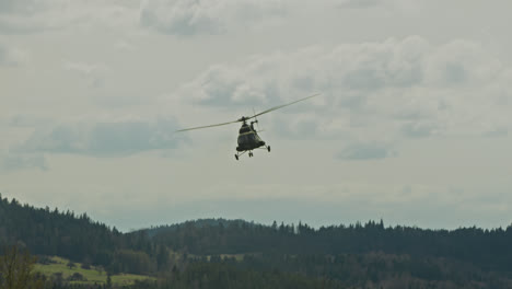 a military-style helicopter flies over dense coniferous forest with overcast skies above, creating a dynamic contrast between nature and machinery. captured in a mountainous region during a day.