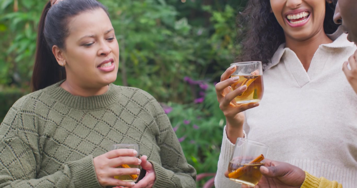 Donne afroamericane che tengono un bicchiere con bastoncini di cannella, controllando la bevanda in giardino mentre un'amica gliela offre.