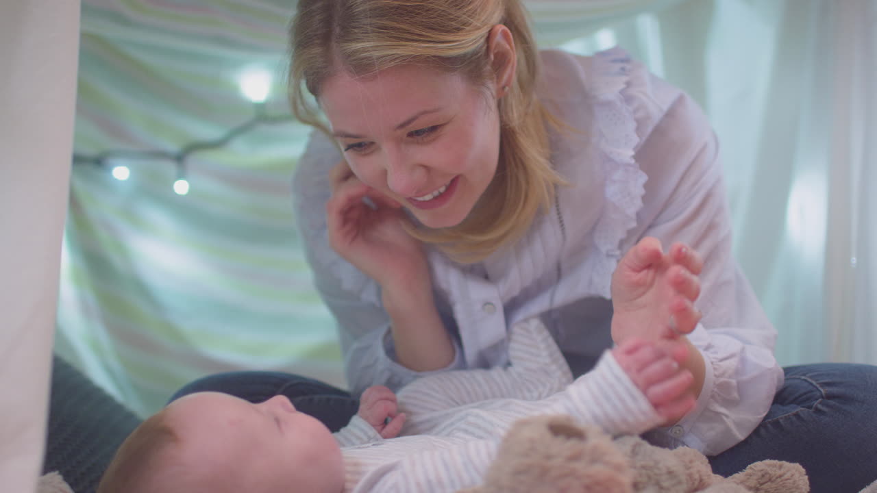 Loving mother playing peek-a-boo with  baby son lying on rug in homemade camp in child's bedroom at home - shot in slow motion