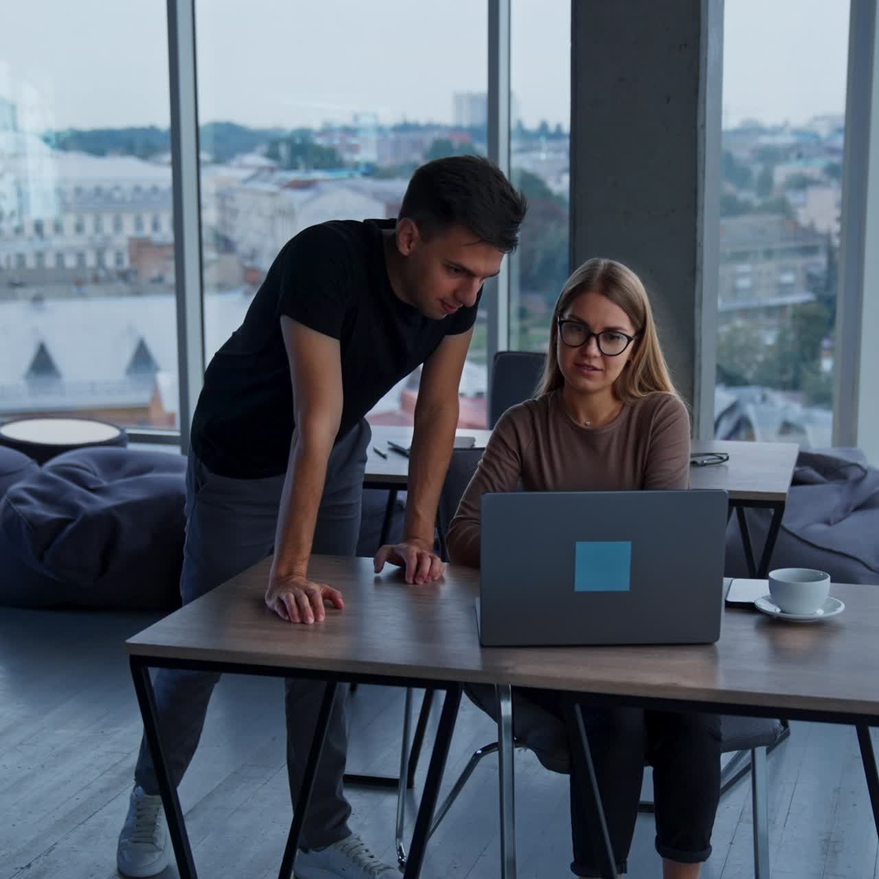 Ufficio moderno, ampio e spazioso con finestre panoramiche e vista sulla città. Persone sedute alle scrivanie che lavorano ai laptop, un uomo si avvicina a una donna per aiutarla