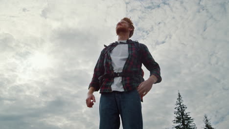 Male-hiker-standing-against-cloudy-sky.-Screaming-man-raising-hands-in-air