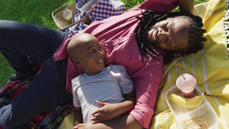 Video-of-happy-african-american-father-and-son-having-picnic-on-grass-and-resting-on-blanket