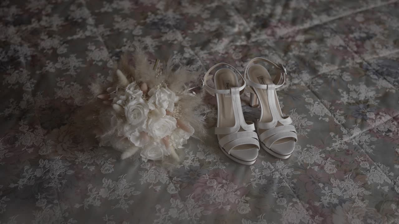 Elegant white bridal shoes and a delicate wedding bouquet placed on floral fabric