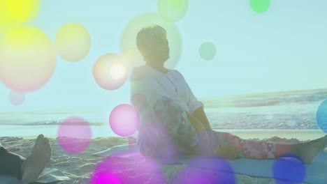 senior woman sitting cross-legged on mat at beach, showing health yoga flow with lens flares