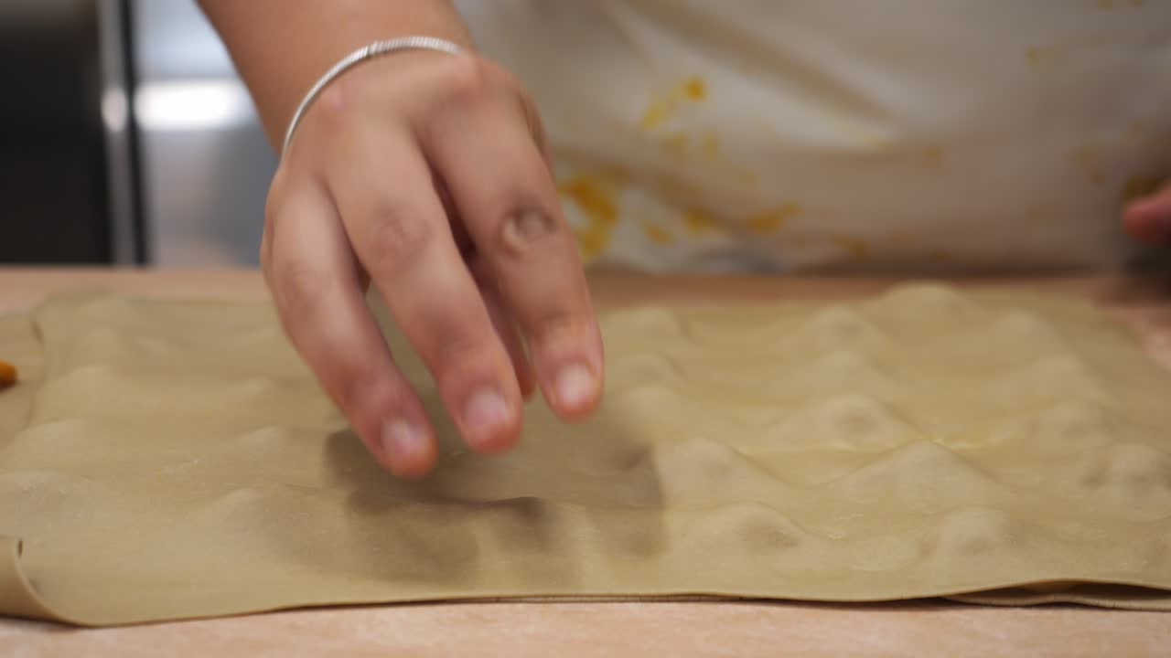 A woman chef places a sheet of pasta over a grid of ravioli filling, pressing gently to seal each pocket before cutting—capturing the precision and rhythm of traditional pasta-making