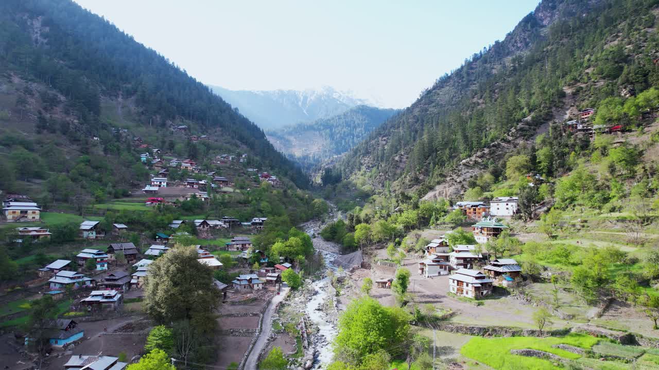 Beautiful view of the Himalayas snowy peaks in the background and a beautiful valley with a stream, forest, green fields, rooftops, and road of Kashmir