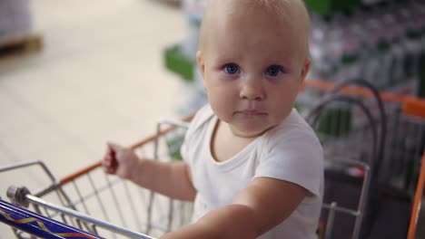 Little-baby-sitting-in-a-grocery-cart-in-a-supermarket,-waiting-for-her-parents-to-do-shopping.-Family-shopping-with-a-little