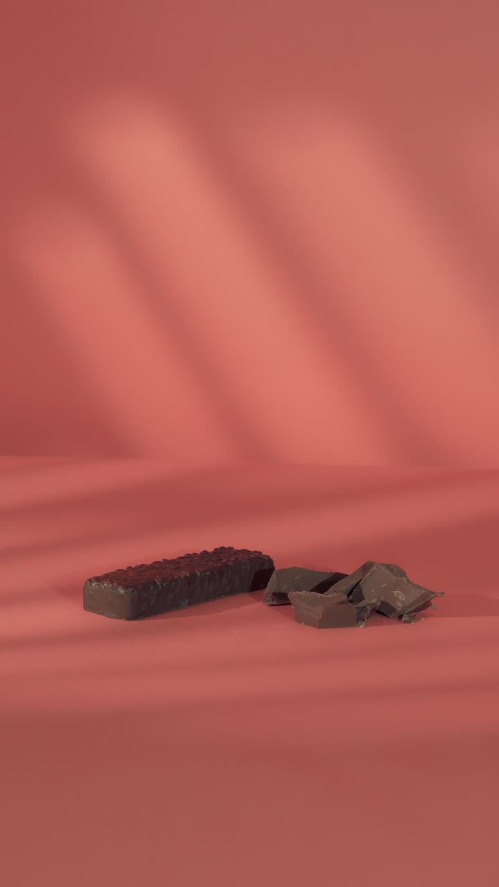 A close-up of a hand placing a dark chocolate-coated protein bar next to broken milk chocolate pieces on a soft coral pink background, highlighting a clean, minimal fitness aesthetic.