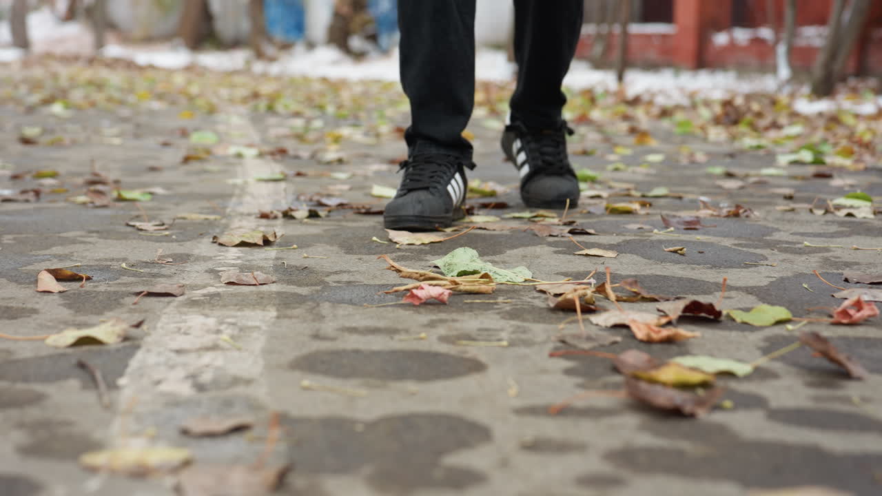 Vue des jambes d'une personne en pantalon noir et baskets marchant le long d'un chemin couvert de feuilles mortes. L'arrière-plan montre des bâtiments et des arbres, créant un environnement urbain naturel.