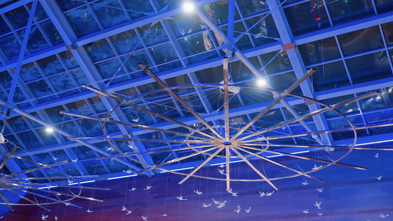 Artistic view of modern mall ceiling with illuminated circular installation featuring wooden spokes and decorative hanging birds against glass roof and blue lights