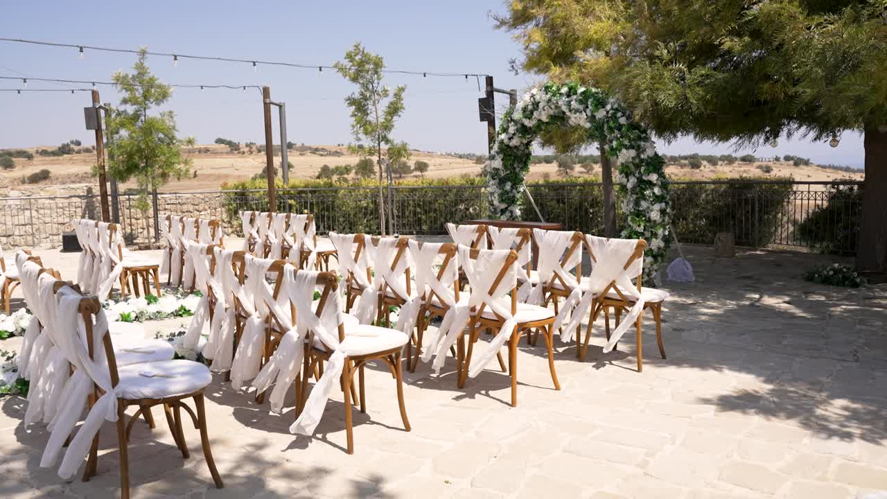 Beautiful outdoor wedding setup in a serene countryside location features white chairs decorated with flowing fabric. Floral archway enhances the charming atmosphere