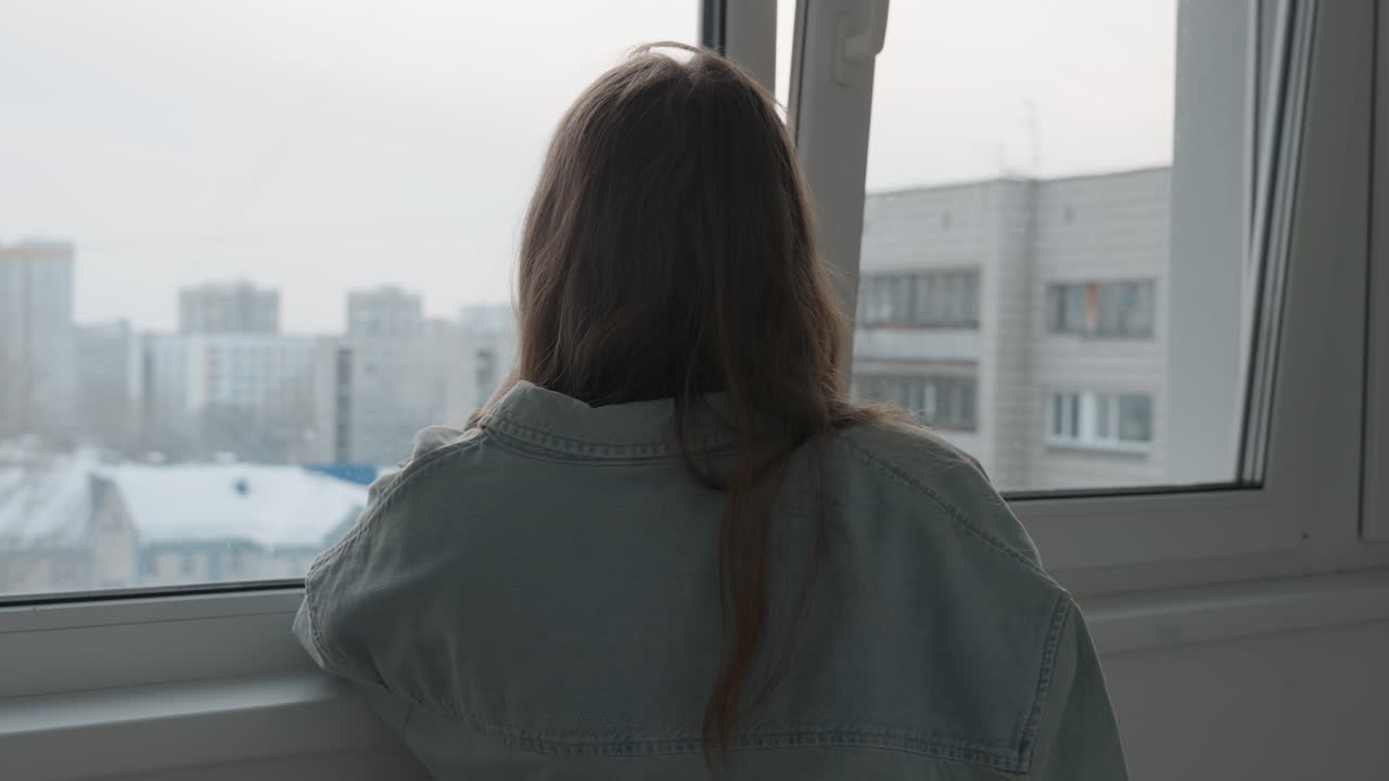 Donna che osserva un paesaggio urbano invernale al chiuso. Una donna che fissa attraverso una finestra ghiacciata una scena cittadina invernale. Una signora che contempla un paesaggio urbano innevato dal comfort dell'interno del suo appartamento