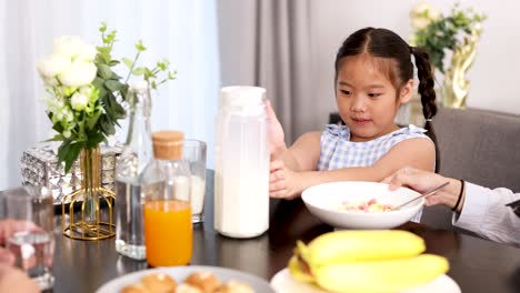 asian child pours milk onto cereal at a bright breakfast table, surrounded by fruit, juice, and family, in a warmly lit modern home