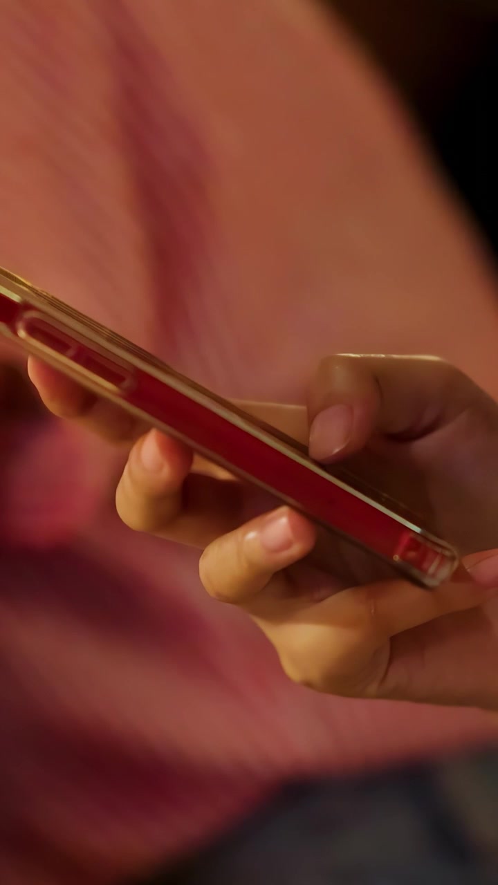 Primo piano di una mano che scorre su uno smartphone rosso, illuminata da una luce soffusa e calda, con uno sfondo sfocato che suggerisce un ambiente interno informale