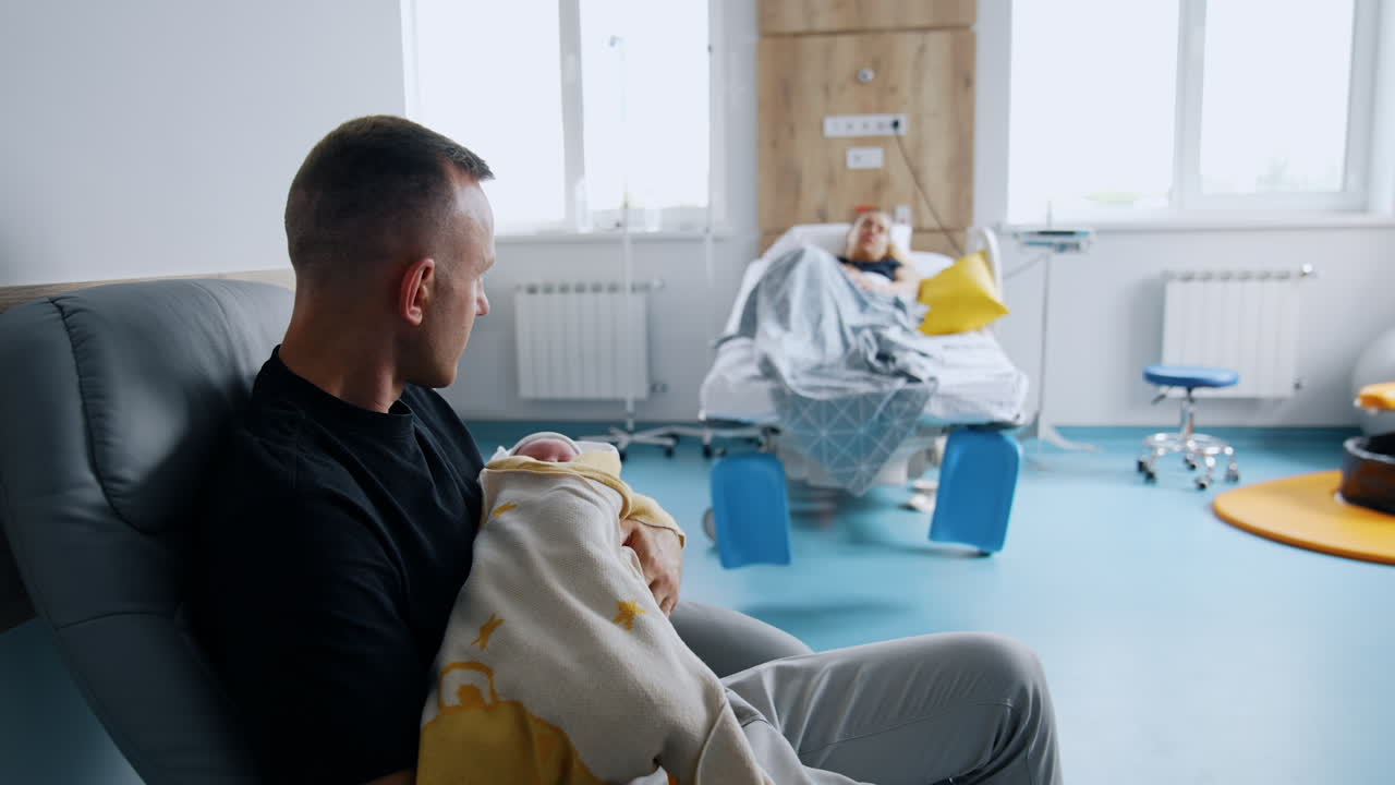 Father is holding a newborn sleeping baby in the hospital ward. Woman is resting in bed at backdrop in blur.