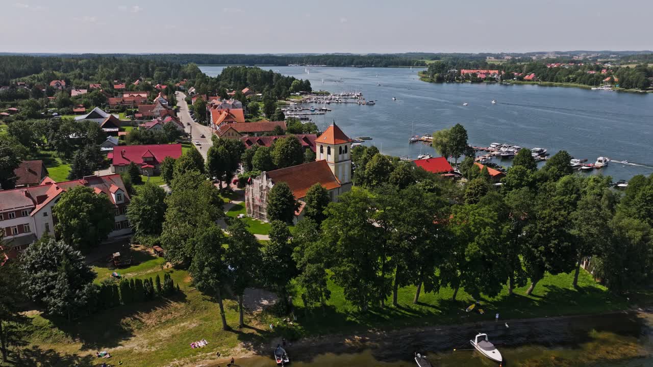 Aerial orbit reveals Rydzewo church green shoreline boats and warm summer views