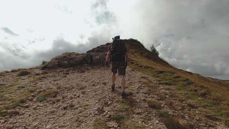 Vista-En-Tercera-Persona-De-Un-Hombre-Caminando-Por-Un-Camino-Rocoso-Hacia-Un-Pico-De-Montaña-Con-Nubes-De-Tormenta-En-El-Horizonte