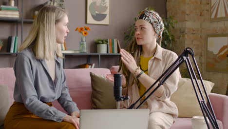 two women recording a podcast talking into a microphone sitting on sofa in front of table with laptop 1