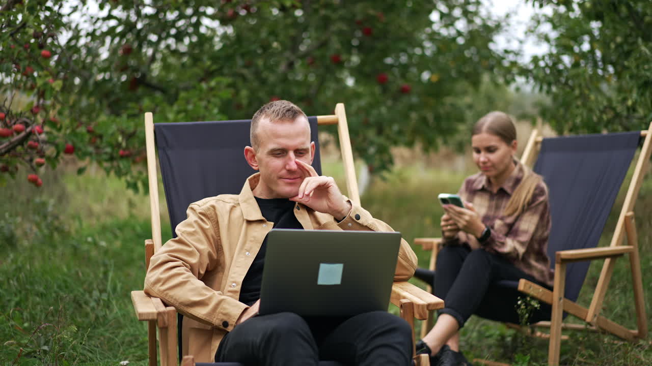 Positive mid aged man working remotely on his laptop. Lady sitting next in the garden chair looks at smartphone in her hands. Apple orchard background.