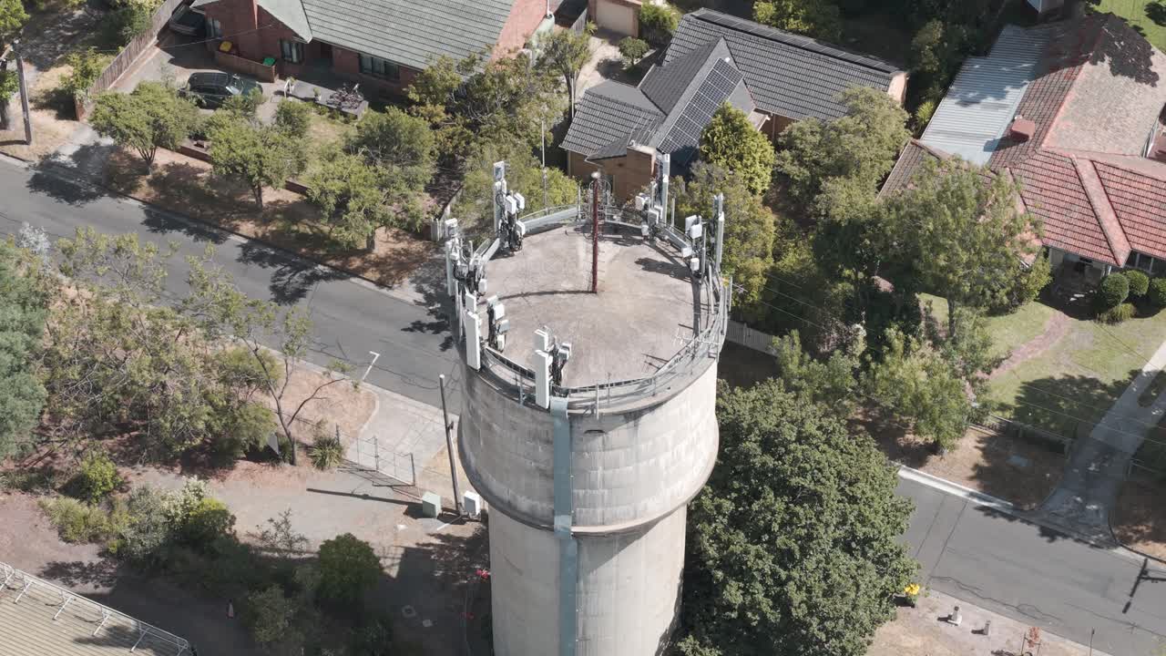 Drohnenvideo zeigt eine aufragende Wasserstruktur, ausgestattet mit 5G-Antennen, inmitten des vorstädtischen Melbourne. Helles Mittagslicht hebt die Stadtlandschaft mit von Bäumen gesäumten Straßen und gemütlichen Häusern hervor.