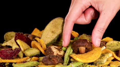 a hand reaches into a colorful pile of assorted dried vegetable chips, picking up a piece. studio lighting highlights textures and vibrant colors, with a static camera and black backdrop