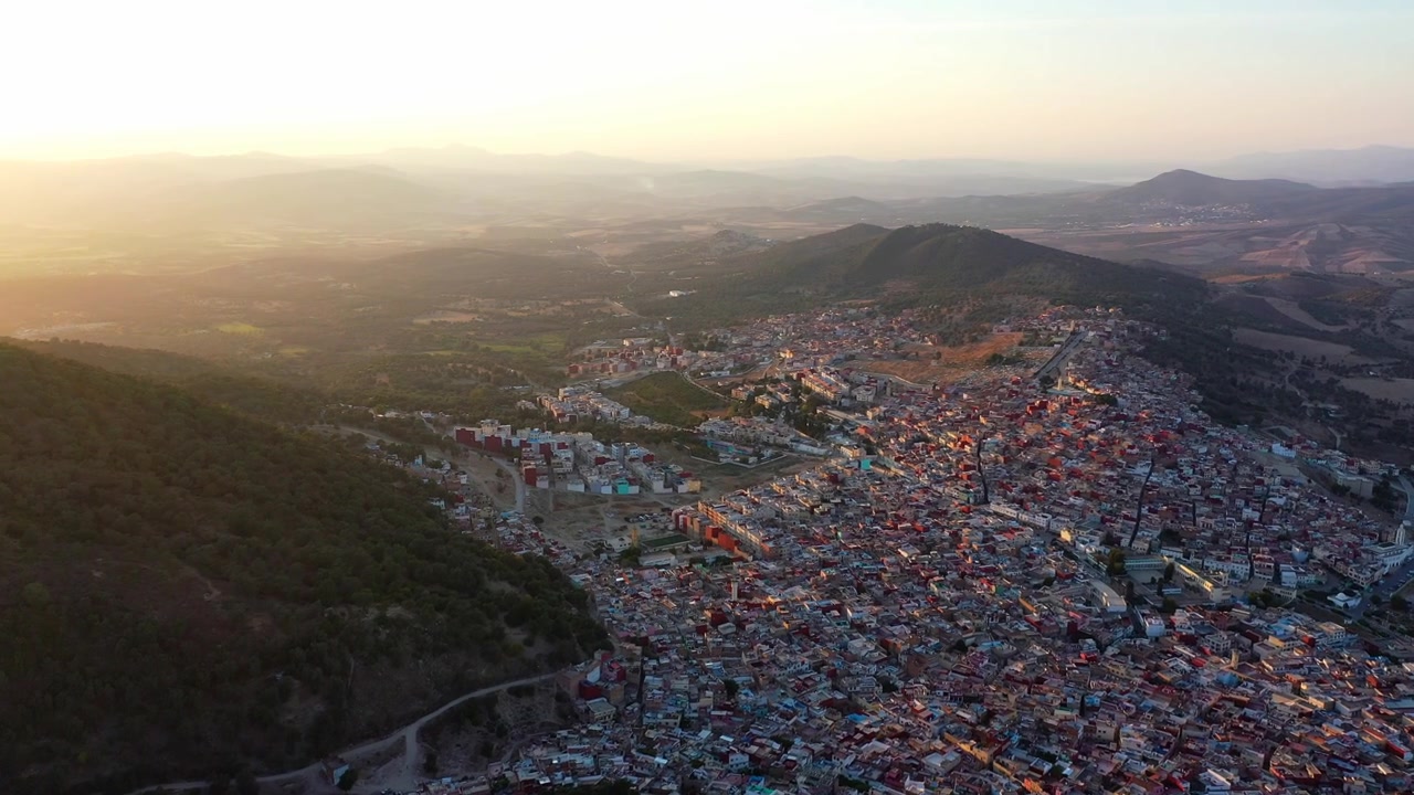 Vista aérea cinematográfica de drone de uma cidade montanhosa marroquina ao pôr do sol, com edifícios urbanos densos, colinas circundantes e luz dourada quente. Ideal para projetos de viagem e documentários.
