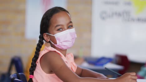 Portrait-of-mixed-race-schoolgirl-wearing-face-mask,-sitting-in-classroom-looking-at-camera