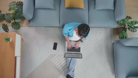 top view of a tired man having a headache while typing on a laptop on the floor with smartphone in front of a sofa at home