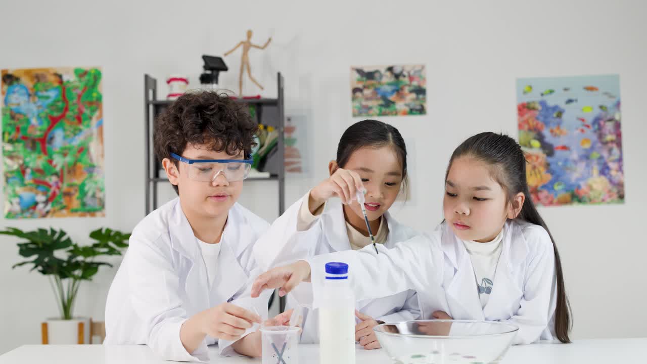 実験着とゴーグルを着用した3人のアジア人の子供たちが、教室でカラフルな化学実験を行っている。