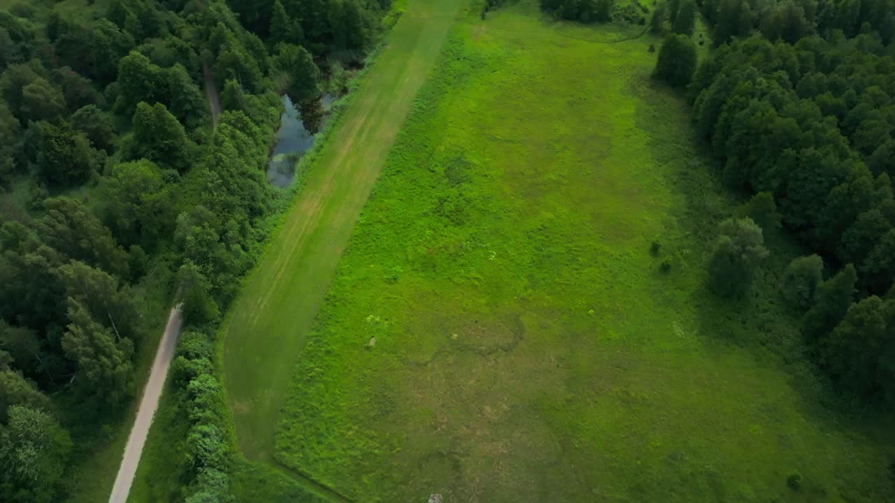 Flying over a rural landscape with an airfield, trees, country road and ponds of water. Downwards tilted view.