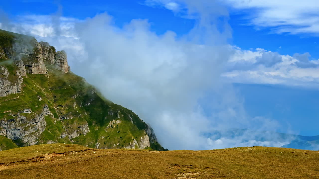 Vista sulle montagne con nuvole ed erba sotto un cielo blu in estate. Degli scalatori si trovano su un pendio erboso mentre le nuvole passano sopra una catena montuosa