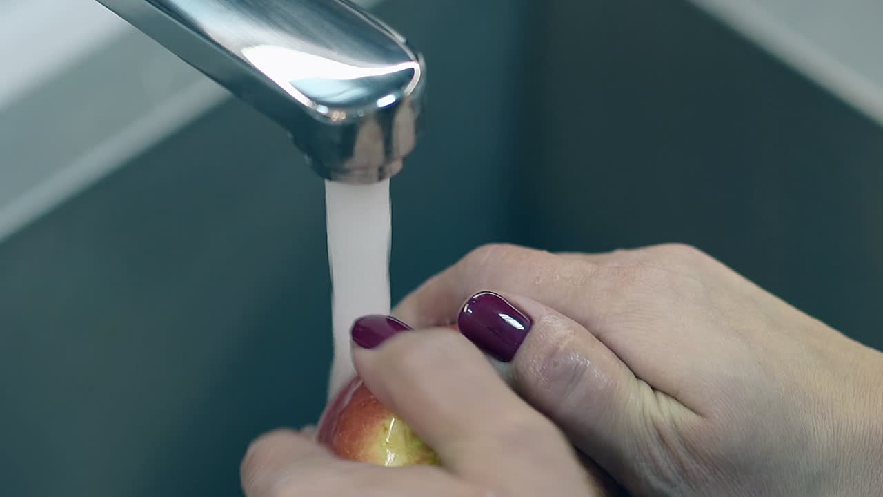 mujer abre el grifo de agua caliente y comienza a lavar gran manzana madura