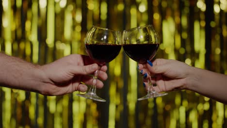 two hands with glasses of red wine making cheers, raising toast on gold background in slow motion