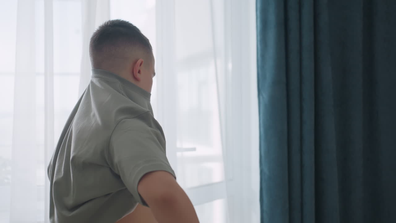 Side view of boy putting on cloth near window with sunlight filtering through curtain capturing simple lifestyle moment of daily dressing routine showing natural indoor activity