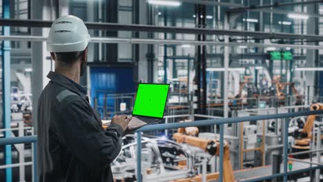 car factory engineer in work uniform using laptop computer with green screen mockup display. working with software at automotive industrial manufacturing facility dedicated for vehicle production.