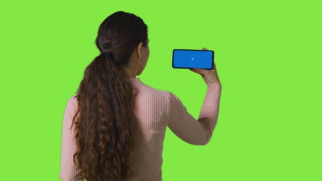 Studio-Rückansicht-Einer-Frau,-Die-Auf-Einem-Bluescreen-Handy-Vor-Einem-Greenscreen-Streamt