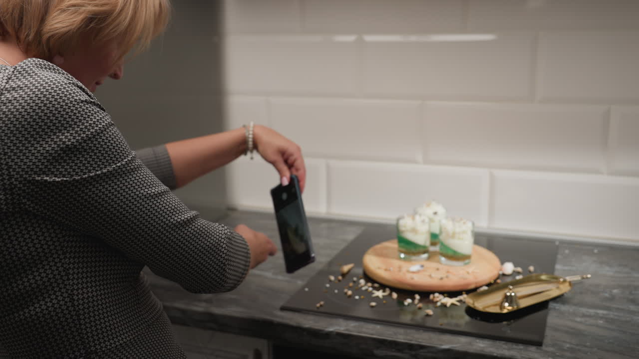 Culinary artist leans forward in kitchen capturing decorated cake candles on wooden tray using smartphone, surrounded by scattered shells and golden utensils in cozy tiled cooking space