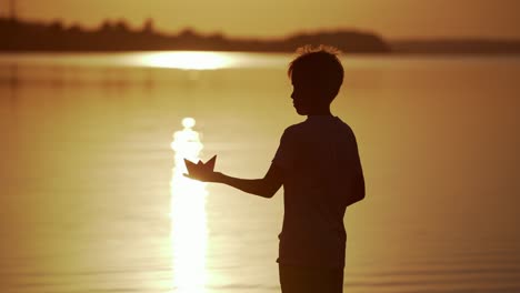 boy with red and green paper ships is standing in the river. origami paper boat in kid's hands. beautiful waves ripple background. travel and nature concept. video at sunset.