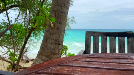 mesa y silla de madera rústica junto a la playa tropical y un árbol con olas turquesas rompiendo en el fondo