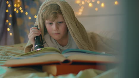 kid reading book with flashlight in bed